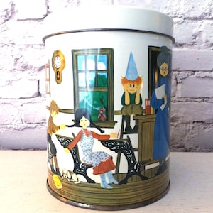 May include: A white metal tin with a lid, featuring a colorful illustration of a classroom scene. The artwork depicts children, a teacher, and a window with a view. A clock hangs on the wall. The tin has a vintage, nostalgic aesthetic.