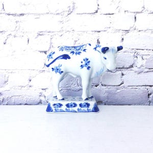 May include: A white ceramic cow figurine with blue floral patterns. The cow stands on a rectangular base, also decorated with blue flowers. The figurine has a classic, decorative style.