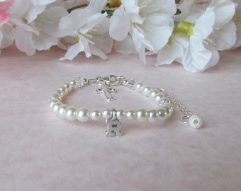 May include: A pearl bracelet with a silver cross charm and a silver letter "R" charm. The bracelet is made of small, round, white pearls and has a silver clasp. The background features white flowers.