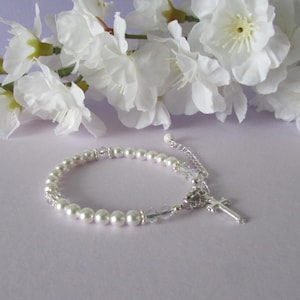May include: A white pearl bracelet with a silver cross charm and clear crystal beads. The bracelet is on a light purple background.