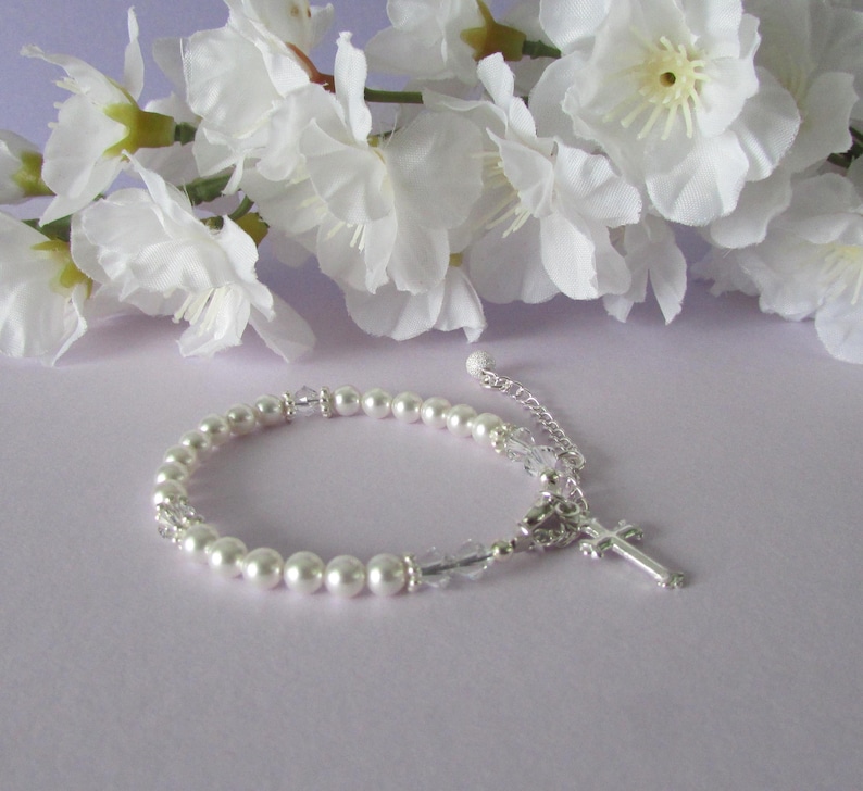 May include: A white pearl bracelet with a silver cross charm and clear crystal beads. The bracelet is on a light purple background.
