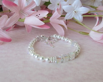 May include: A white pearl and crystal bracelet with a silver cross charm. The bracelet is on a pink background with white flowers.