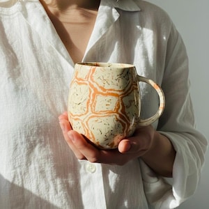 Könnte beinhalten: Eine Keramik-Tasse mit einem einzigartigen Muster aus orangefarbenen und schwarzen Wirbeln auf cremefarbenem Hintergrund. Die Tasse hat eine abgerundete Form und einen gebogenen Henkel. Die Tasse wird in den Händen einer Person gehalten, vor einem weißen Hemd.