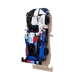 May include: A Lego model car in blue, white, red, and black, with visible wheels and the word "Motorsport" on the front. The car is made from Lego bricks and has a streamlined design. The model is approximately 30 cm in length.