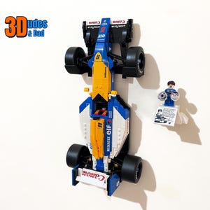 May include: A blue, yellow, and white Lego race car model with black wheels. The car has the Canon and Elf logos. A Lego figurine is holding two silver wheels. The text "3Dudes & Dad" is in the upper left corner.