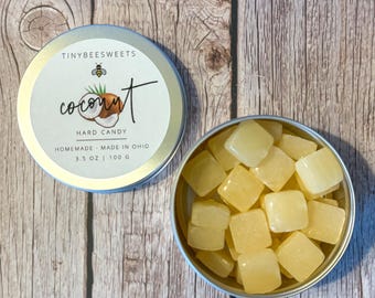Puede incluir: Una lata de plata con una etiqueta blanca que dice "Tinybee Sweets Coconut Hard Candy Homemade - Made in Ohio 3.5 oz | 100 g". La lata está abierta y llena de pequeños caramelos de coco cuadrados y amarillos.