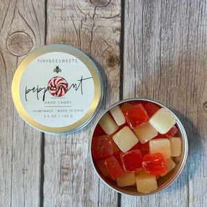 May include: A round tin with a gold lid and white label that reads "TINYBEESWEETS peppermint HARD CANDY HOMEMADE - MADE IN OHIO 3.5 OZ | 100 G". The tin is filled with red and white square hard candies.