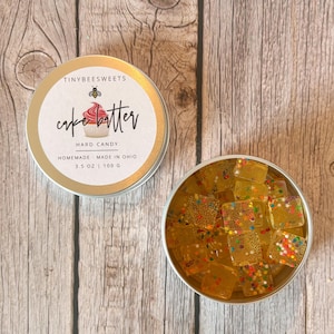 May include: Two round metal tins on a wooden surface. One tin is closed, displaying the text "cake batter" and a cupcake graphic. The other tin is open, filled with clear, square hard candies with colorful sprinkles. The text on the closed tin reads "Hard Candy, Homemade - Made in Ohio, 3.5 oz | 100 G."