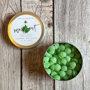 May include: Open tin of green sweet mint breath mints next to a tin with a label that reads "Tiny Bee Sweets" and "sweet mint" with a bee graphic. The mints are homemade in Ohio and the tin contains 3.5 oz or 100 g.
