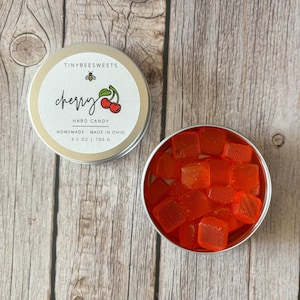 May include: A silver tin with a white label that reads "TINYBEESWEETS cherry HARD CANDY HOMEMADE - MADE IN OHIO 3.5 OZ | 100 G". A small bee illustration is on the label. The tin is filled with orange-red square hard candies.