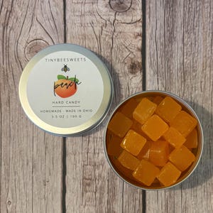 May include: A silver tin filled with orange hard candy. The tin has a label that says "Tinybee Sweets" and "Peach Hard Candy" with a small bee illustration. The label also says "Homemade - Made in Ohio" and "3.5 oz | 100 g".