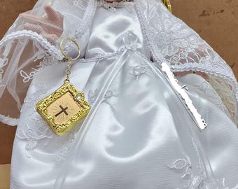 Könnte beinhalten: Eine weiße Satin-Babypuppe, die ein weißes Spitzenkleid mit einem goldenen Kreuz-Schlüsselanhänger trägt. Die Puppe sitzt auf einem goldenen Thron mit Engelsflügeln.