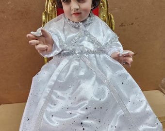 Könnte beinhalten: Detaillierte Statue einer Kinderfigur, bekleidet mit einem weißen Satinkleid mit silbernen Verzierungen und passendem Kopfschmuck. Die Figur sitzt in einem goldenen Stuhl mit rotem Rücken. Der Text "Niño de las Palomitas" steht unten.