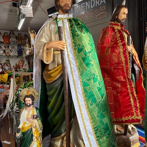 Op de afbeelding: Beelden van religieuze figuren, waaronder een met een groene en gouden cape en een staf, en een andere in een rode cape. De beelden staan in een winkel met een bord met de tekst "ALMENDRA" en andere religieuze beeldjes op de achtergrond.