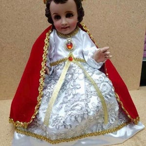 May include: A white and silver dress with gold trim and a red cape. The doll is wearing a gold crown with red jewels. The doll has brown hair and a painted face. The doll is sitting on a white cushion. The text "SAGRADO CORAZÓN" is printed below the doll.
