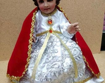 Könnte beinhalten: Ein weißes und silbernes Kleid mit goldenen Verzierungen und einem roten Umhang. Die Puppe trägt eine goldene Krone mit roten Juwelen. Die Puppe hat braunes Haar und ein bemaltes Gesicht. Die Puppe sitzt auf einem weißen Kissen. Der Text "SAGRADO CORAZÓN" ist unter der Puppe gedruckt.