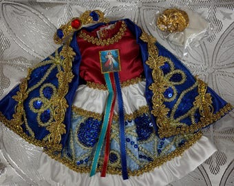 Puede incluir: Un vestido de muñeca blanco, azul y dorado con una faja roja y una corona. El vestido tiene una capa azul con ribetes dorados y lentejuelas. La corona es dorada con joyas rojas. El vestido de muñeca es para una figura religiosa.