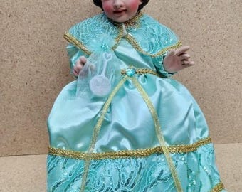 Puede incluir: Una muñeca vestida con un vestido de satén turquesa y encaje con ribetes dorados. La muñeca lleva una corona con una gema turquesa. La muñeca sostiene un juguete de plástico blanco.
