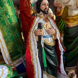 Puede incluir: Estatua religiosa de un hombre con cabello oscuro y barba, vistiendo una capa roja y blanca con adornos dorados. Sostiene un bastón y un medallón. Tela verde y dorada en el fondo.