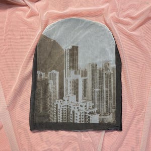 May include: A rectangular piece of sheer fabric with a cityscape print, showing high-rise buildings and a mountain, set against a pink backdrop. The fabric has a dark border, possibly for a decorative item or part of a garment.