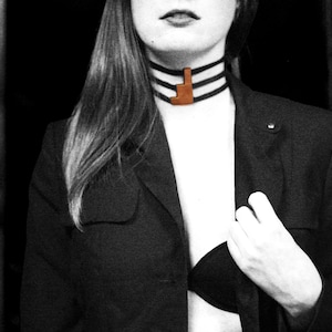 Op de afbeelding: Zwart-wit afbeelding van een persoon die een zwarte blazer draagt en een zwarte choker-ketting met drie strengen en een bruine hanger. De persoon draagt een zwarte bh. De achtergrond is zwart.