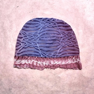 May include: A beanie hat featuring a textured, ruched fabric design. The upper portion is a gradient of purple and blue, while the lower band is a contrasting reddish-pink hue. The material has a semi-sheer, layered appearance with horizontal lines.