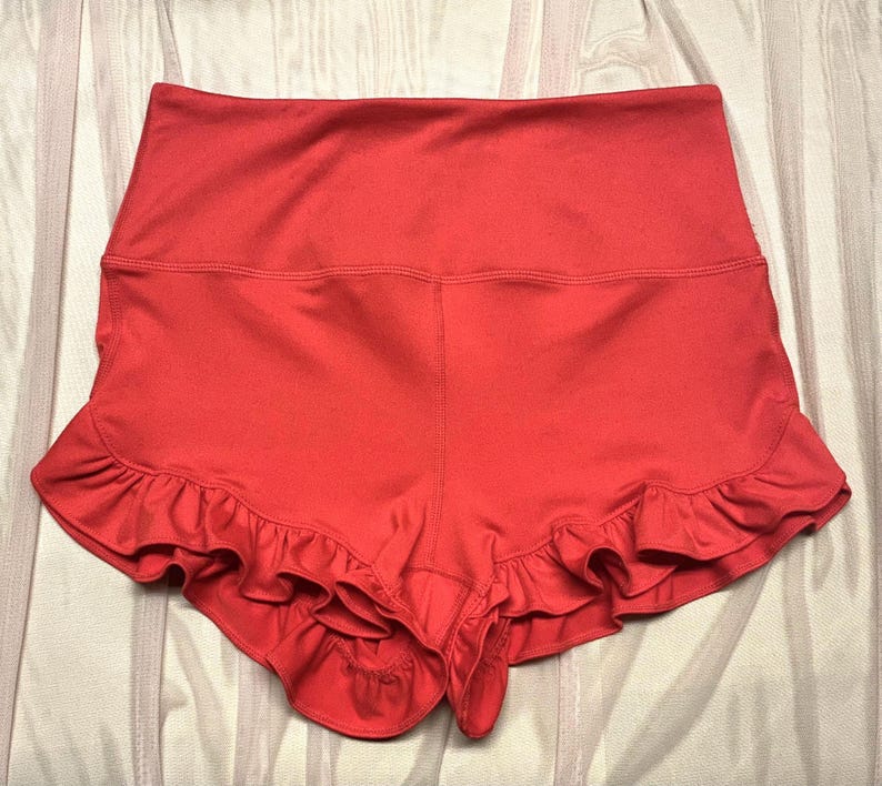 May include: Coral-colored athletic shorts with a high waistband and a ruffled hem. The shorts are made of a smooth, stretchy fabric and are designed for comfort and movement. The ruffled hem adds a feminine touch to the sporty design.