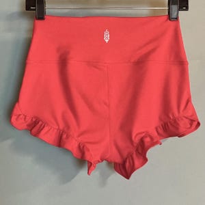 May include: Coral-colored shorts with a high waistband and a ruffled hem. A white floral logo is centered on the waistband. The shorts are hanging from a black hanger.
