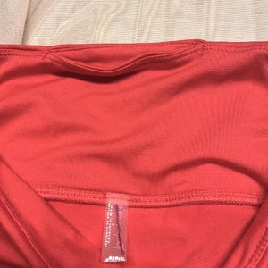 May include: Close-up of a coral-colored athletic top. The top features a scoop neckline and a small pocket detail at the back. A tag with text is visible at the bottom. The fabric appears to be a lightweight, breathable material.