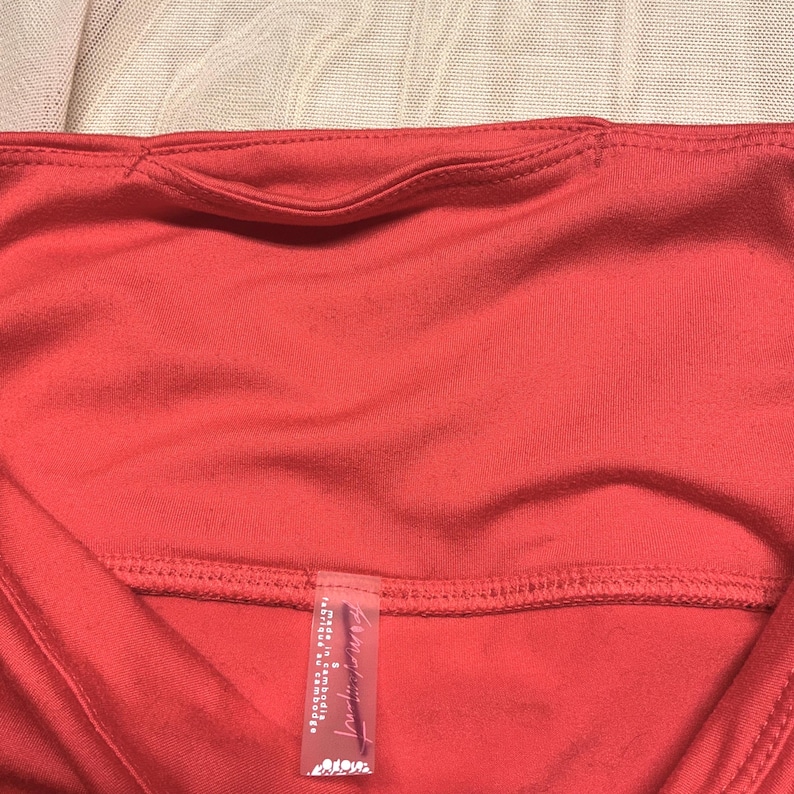 May include: Close-up of a coral-colored athletic top. The top features a scoop neckline and a small pocket detail at the back. A tag with text is visible at the bottom. The fabric appears to be a lightweight, breathable material.