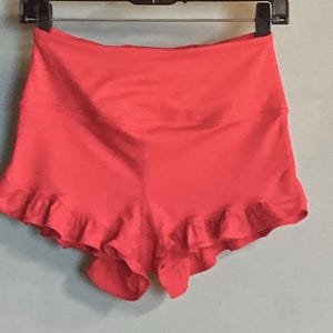 May include: Coral-colored shorts with a high waistband and a ruffled hem. The shorts are hanging on a black hanger against a light blue wall. The shorts appear to be made of a smooth, stretchy fabric.