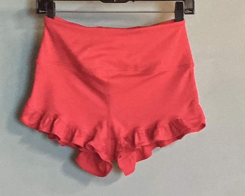 May include: Coral-colored shorts with a high waistband and a ruffled hem. The shorts are hanging on a black hanger against a light blue wall. The shorts appear to be made of a smooth, stretchy fabric.
