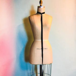 May include: A black velvet ribbon choker necklace on a dress form. The dress form is on a metal stand. The background is a gradient of pink and blue.