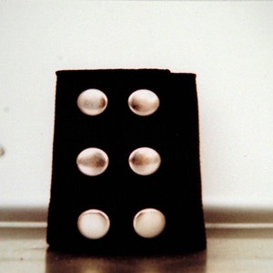 May include: A black leather cuff with six silver-colored, rounded studs. The cuff is rectangular and appears to be made of a soft material. The studs are evenly spaced and have a shiny, metallic appearance.