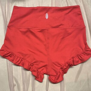 May include: Coral-colored athletic shorts with a high waistband and ruffled hem. A small white logo is centered on the back of the shorts. The shorts are made of a smooth, stretchy material.