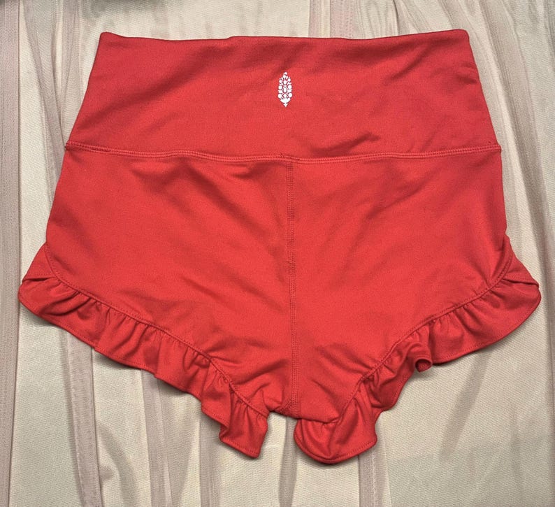 May include: Coral-colored athletic shorts with a high waistband and ruffled hem. A small white logo is centered on the back of the shorts. The shorts are made of a smooth, stretchy material.