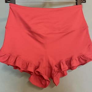 May include: Coral-colored athletic shorts with a high waistband and a ruffled hem. The shorts are made of a smooth, stretchy fabric and are hanging on a black hanger. The shorts are designed for athletic activities.