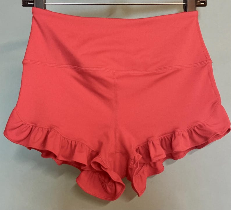 May include: Coral-colored athletic shorts with a high waistband and a ruffled hem. The shorts are made of a smooth, stretchy fabric and are hanging on a black hanger. The shorts are designed for athletic activities.