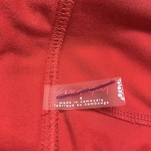 May include: Close-up of a vibrant red garment with visible stitching. A transparent label is attached, displaying the text "from movement" and "made in cambodia". The label also indicates a size "S".