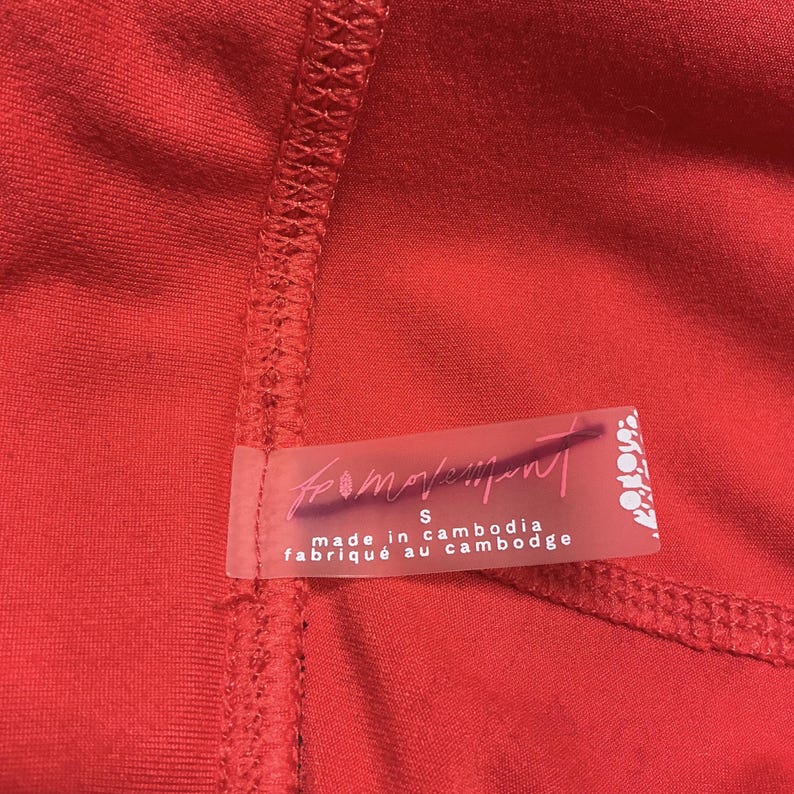 May include: Close-up of a vibrant red garment with visible stitching. A transparent label is attached, displaying the text "from movement" and "made in cambodia". The label also indicates a size "S".