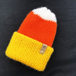 May include: A knitted hat in the shape of a candy corn with yellow, orange, and white stripes. The hat has a brown leather tag with the text "DLK" on it.