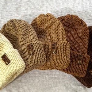 May include: Five knitted beanies in various shades of brown, beige, and cream. Each beanie has a small leather patch with the letters "BLK" and a heart symbol.