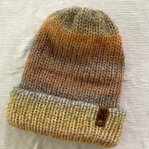 May include: A knitted beanie hat with a striped pattern in shades of orange, brown, and gray. The hat has a fold-over cuff and a small leather patch with a heart symbol.