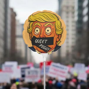 May include: A round balloon on a white stick featuring a cartoon face with yellow hair and a black strip over the mouth with the words "QUIET PIGGY". The balloon is held up in front of a crowd of people.