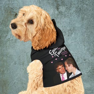 May include: A light brown dog wearing a black hoodie with the text "Epstein and Trump Friends Forever" and images of Donald Trump and another man. The hoodie has pink heart accents.