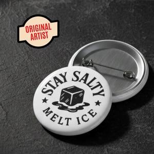 Może przedstawiać: Biała przypinka z napisem "STAY SALTY MELT ICE" w okrągłym wzorze, z grafiką topniejącej kostki lodu. Obok znajduje się otwarty tył przypinki. Obraz zawiera również małe logo "ORIGINAL ARTIST".