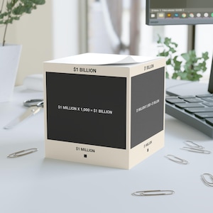 May include: A cube-shaped notepad with the text "$1 BILLION" at the top and "$1 MILLION" at the bottom. The center of the cube has the text "$1 MILLION X 1,000 = $1 BILLION" on a black background. The notepad is on a white surface.