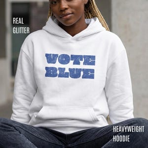 May include: A white hooded sweatshirt featuring the text VOTE BLUE in a distressed blue font across the chest. The garment includes a front kangaroo pocket, long sleeves, and a hood with drawstrings. The text REAL GLITTER and HEAVYWEIGHT HOODIE appear.