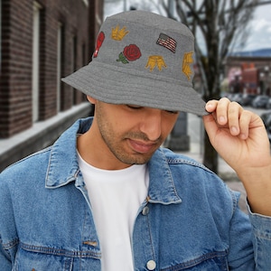 May include: A grey bucket hat featuring embroidered patches including a red rose, crowns, and an American flag. The hat is made of a textured fabric and has a wide brim. The hat is being held by a person.