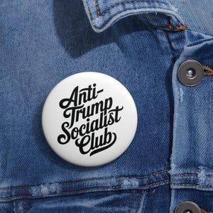 May include: A white button with the text "Anti-Trump Socialist Club" in black script. The button is round and is set against a blue denim jacket.